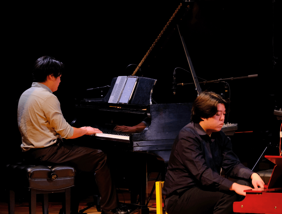 Two pianists perform together on stage, playing side by side on grand pianos during a concert.