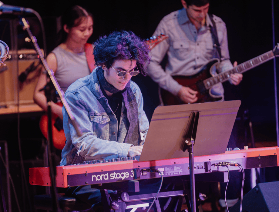 Student musicians perform at the Songwriter Showcase, featuring a keyboardist and guitarists playing on stage under concert lighting.
