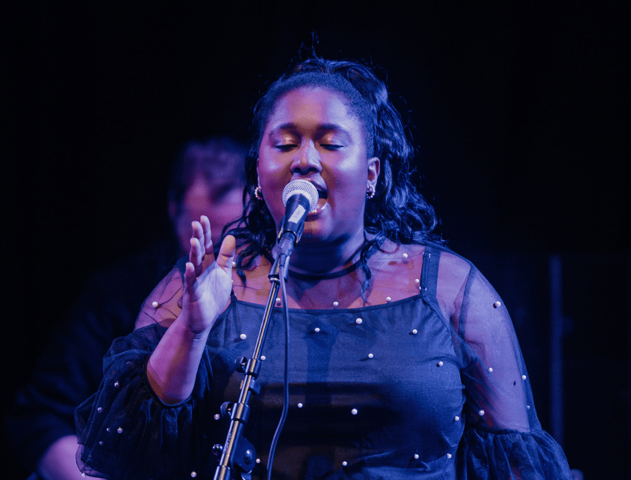 A vocalist performs passionately on stage during the Songwriter Showcase, singing into a microphone under blue stage lighting.