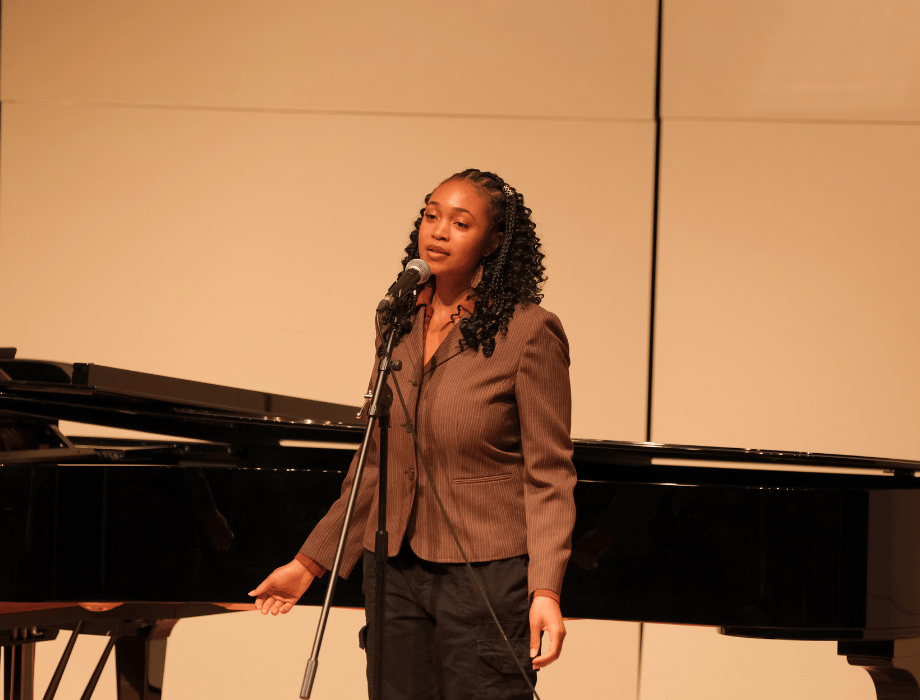 A vocalist performs on stage beside a grand piano, singing into a microphone during a recital performance.