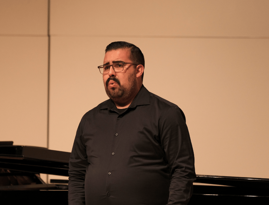 A vocalist performs on stage beside a piano during a recital, singing expressively in a formal setting.