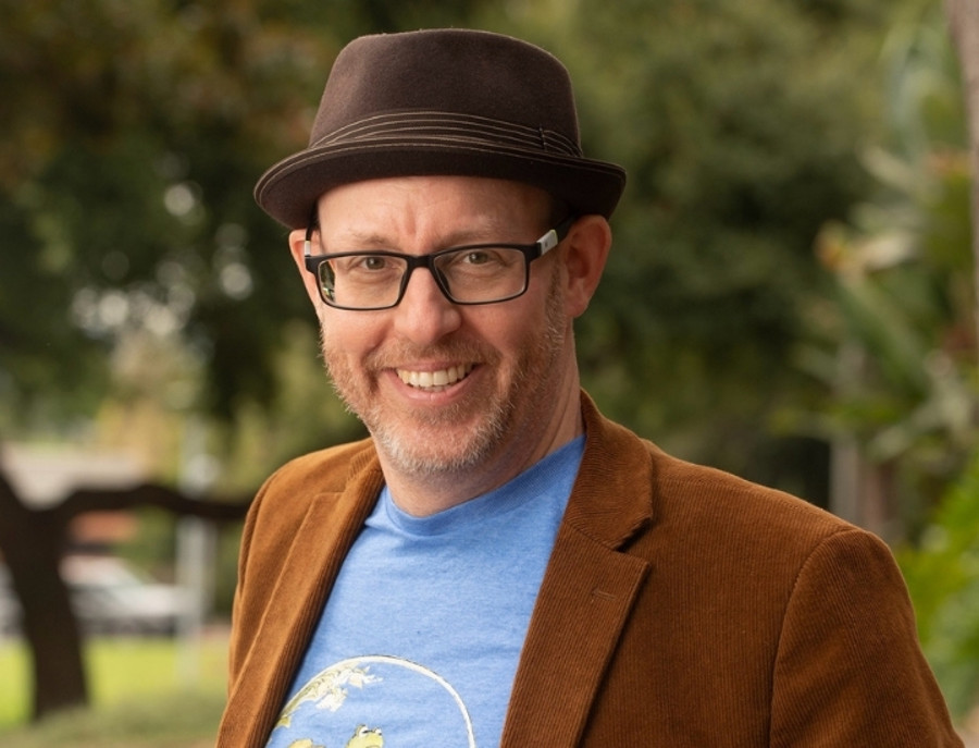 Dr. Evan Ware, Department Chair, smiles outdoors wearing a hat, glasses, and a brown blazer.