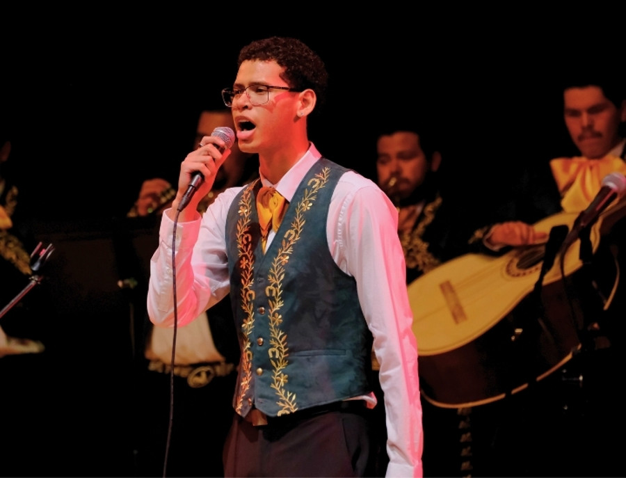 Mariachi vocalist performing on stage with live ensemble and stringed instruments in the background.