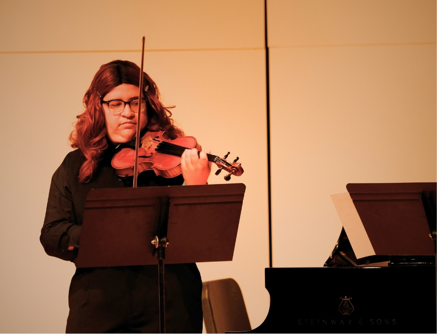 Student violinist performs on stage with focused expression, reading sheet music during a recital.