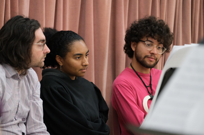 Three music students listen intently during a composition review session with sheet music visible.