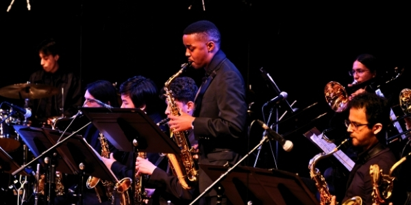 A jazz band performs on stage, featuring a diverse group of musicians in black attire playing saxophones, trombones, and drums. One saxophonist stands in the center, soloing under a spotlight, while others play seated behind music stands.
