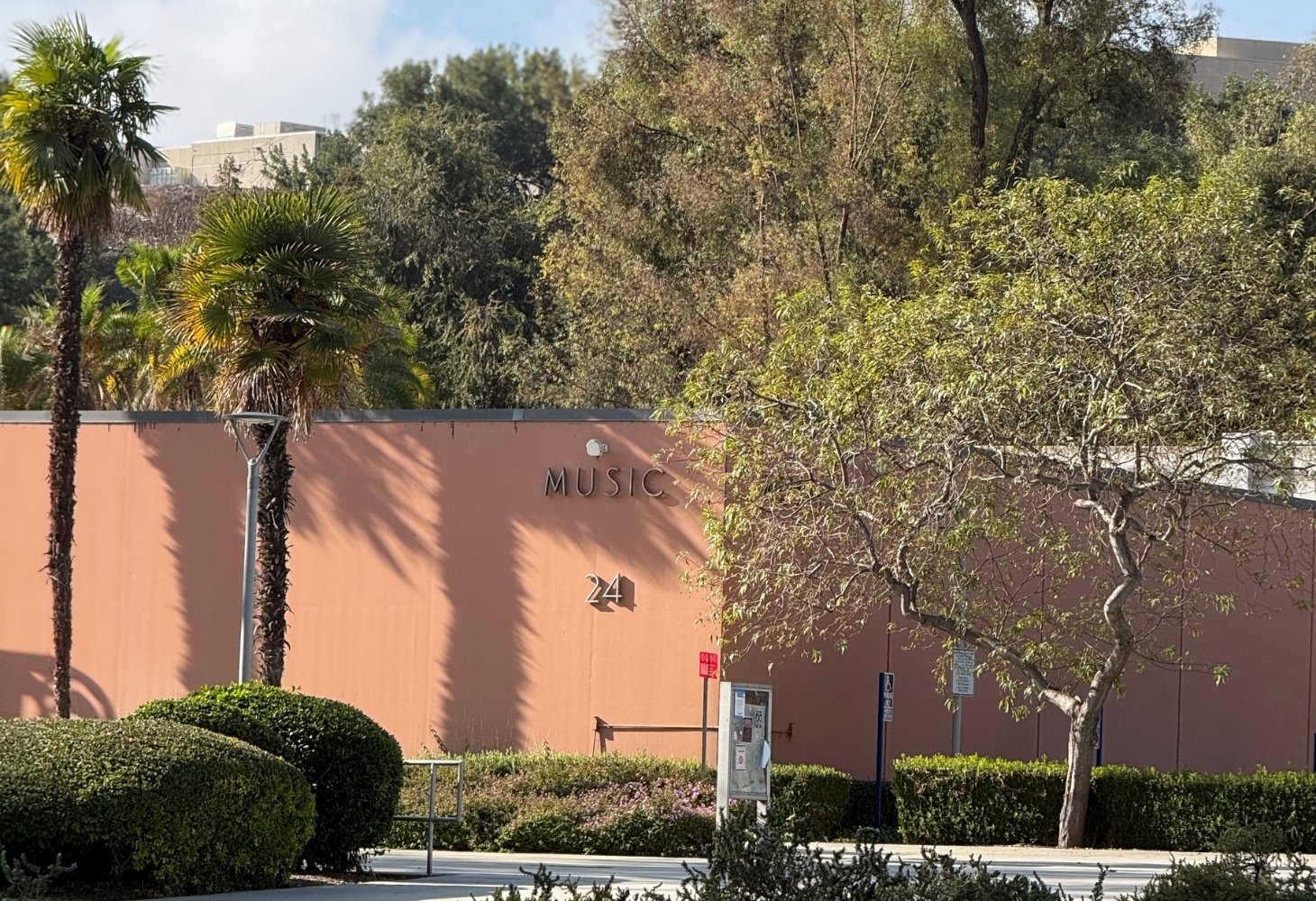 Exterior view of a peach-colored building labeled “MUSIC 24,” partially shaded by trees and surrounded by palm trees.