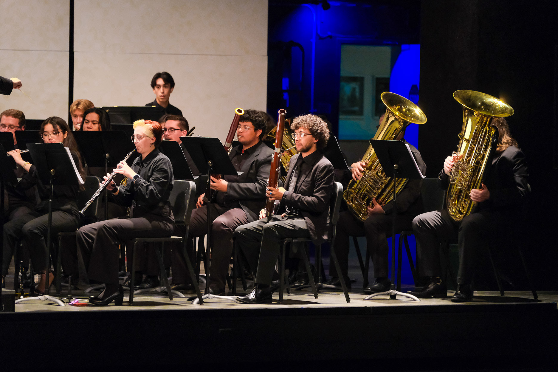 Wind ensemble musicians perform on stage with clarinets, bassoons, and tubas under stage lighting.