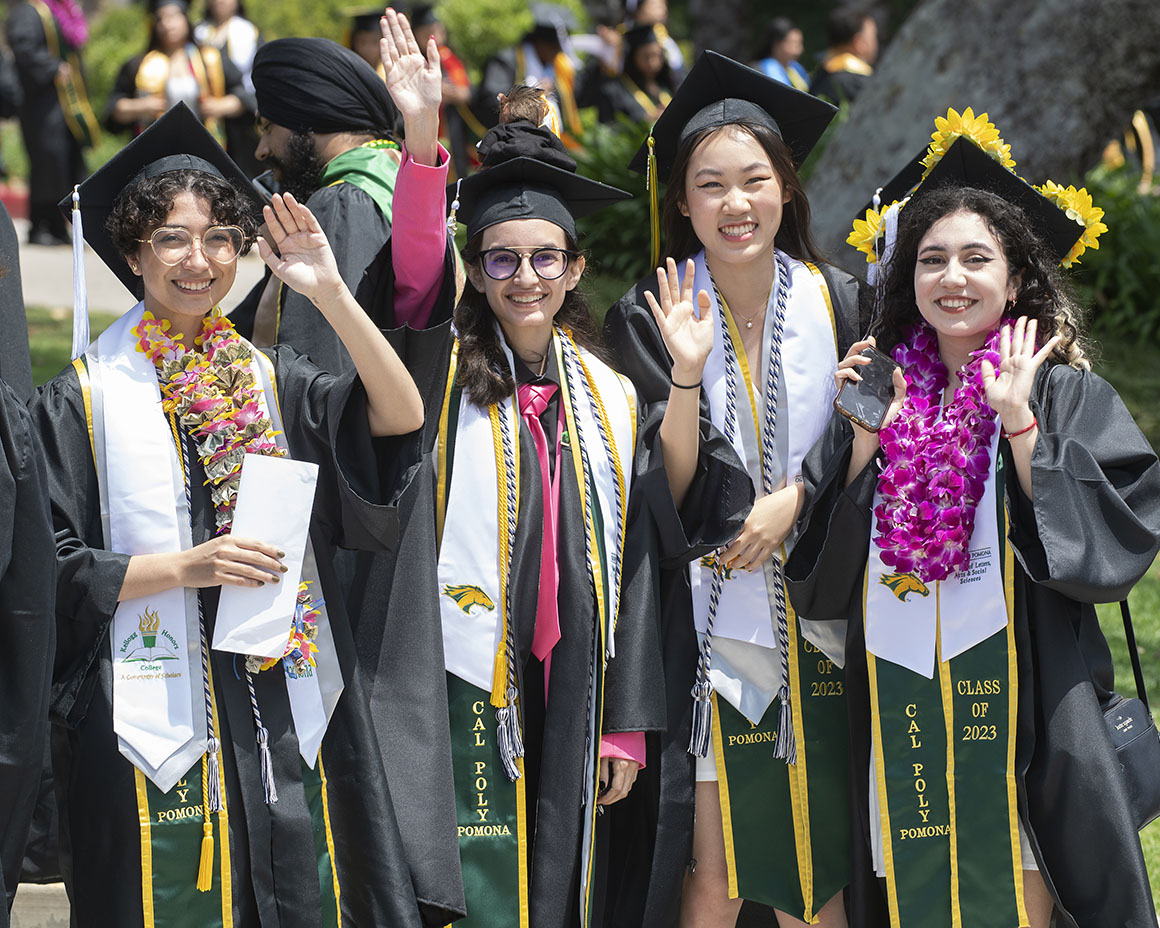 Four Cal Poly Pomona graduates in caps and gowns smile and wave at commencement.