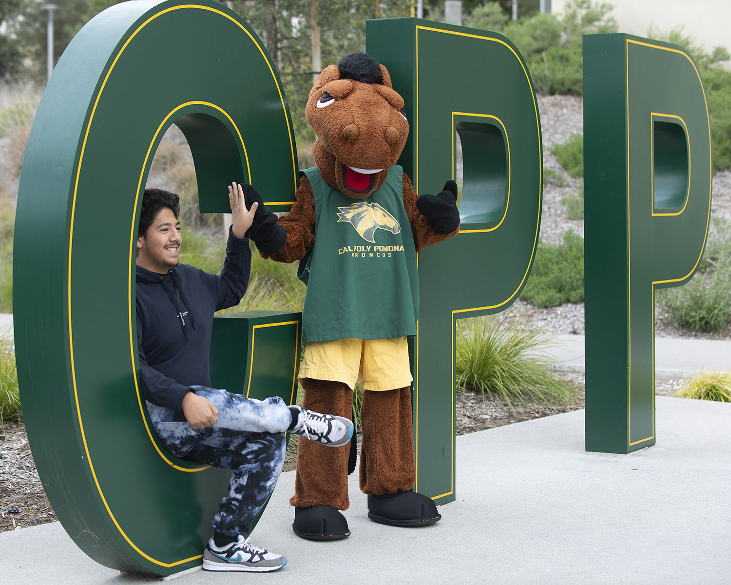 A student high-fives the Cal Poly Pomona Bronco mascot in front of large CPP letters.