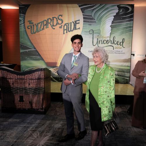 A young man in a gray suit stands beside an older woman wearing a green jacket at a formal event. They pose smiling in front of a decorative backdrop reading “The Wizards of Awe” and “Hospitality Uncorked 2024." 