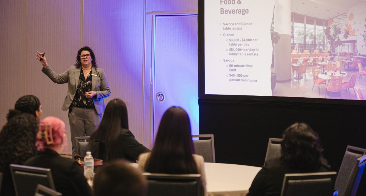 Woman standing inside a hotel ballroom in front of projector and students listening at tables