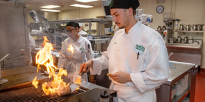 student chef flipping a burger on a grill with flames