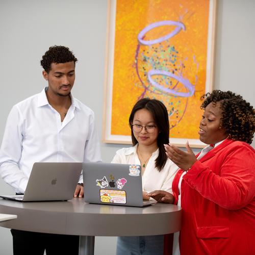 Three people stand around a tall table collaborating on laptops in a modern indoor space. A woman gestures while speaking as a man and another woman look at their screens, conveying teamwork, discussion, and professional collaboration.