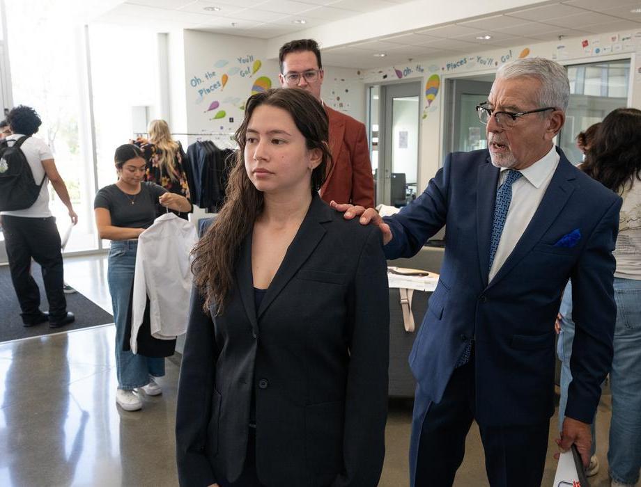 A woman wearing a black blazer stands in a bright indoor space while an older man in a blue suit gently adjusts the shoulder of her jacket. Other people are visible in the background holding clothing, suggesting a professional styling.
