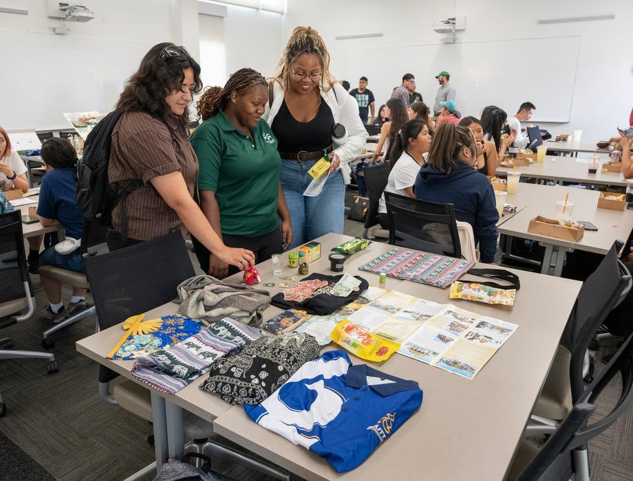 Three people stand around a table covered with folded clothing, snacks, small items, and printed materials during a busy classroom or workshop event. Other participants sit at tables in the background.