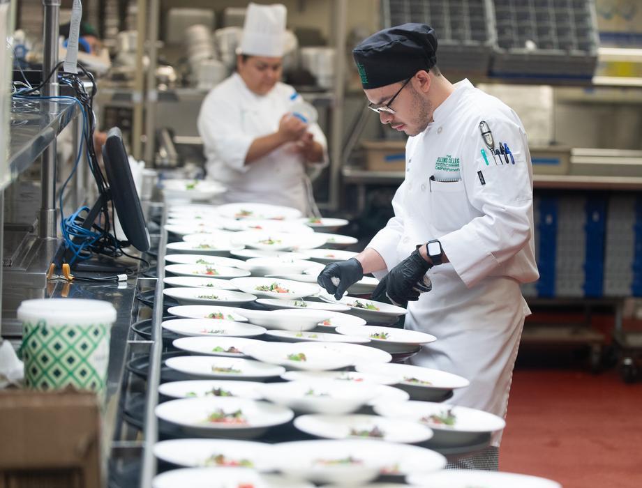 A culinary student wearing a white chef coat and black gloves carefully plates dishes along a stainless steel counter in a commercial kitchen. Rows of prepared plates extend into the background while another chef works behind them, highlighting hands-on culinary training and professional food preparation.