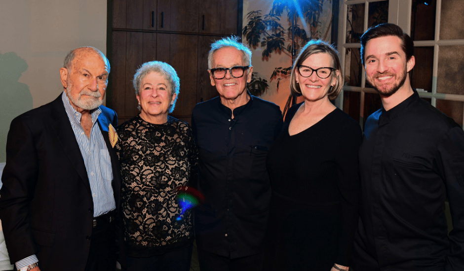 Five adults stand shoulder to shoulder smiling at an indoor event with warm lighting and plants in the background.