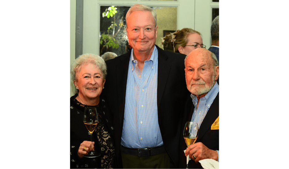 Three adults pose together holding wine glasses at a formal indoor gathering near large windows.
