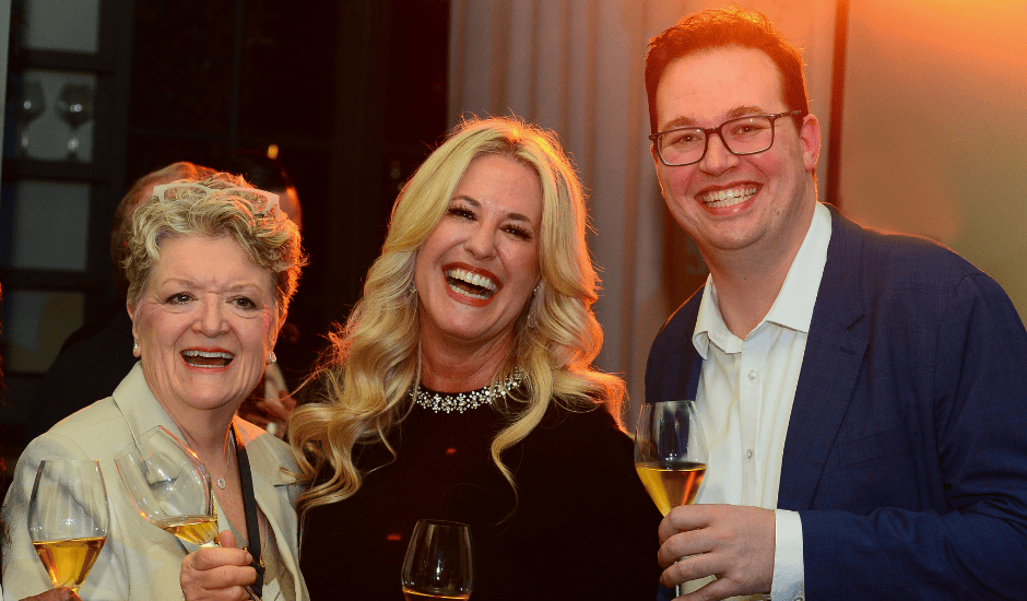 Three adults laugh together while holding wine glasses at a warmly lit indoor gathering.