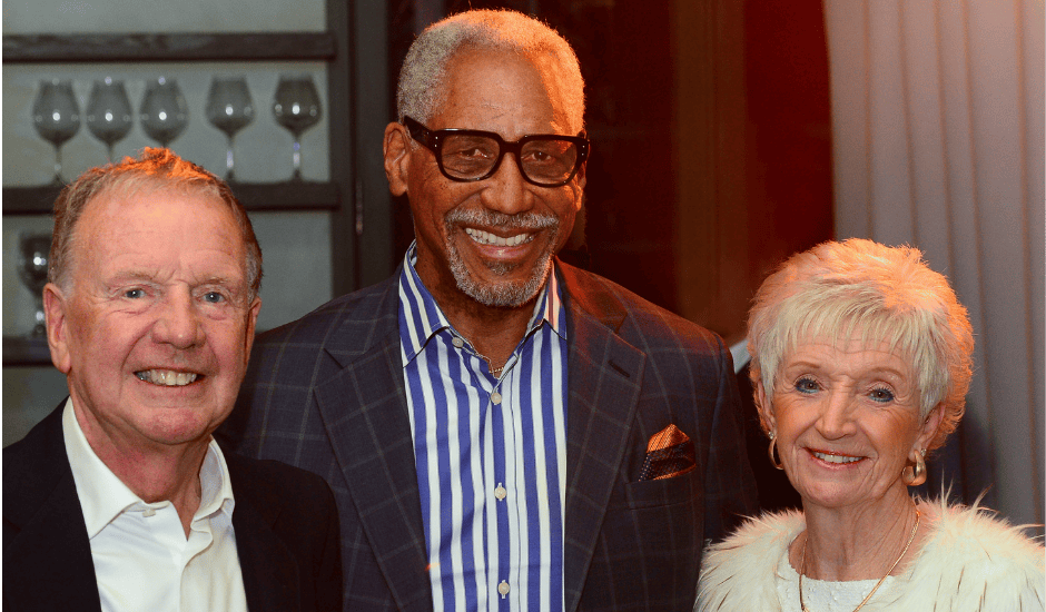 Three adults pose shoulder to shoulder at an evening event with shelves of glassware behind them.