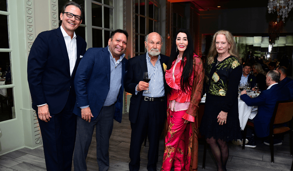 Five adults stand together at a formal indoor dinner with seated guests visible in the background.