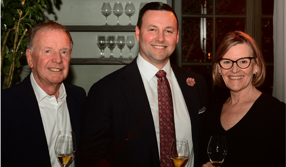Three adults smile while holding wine glasses at an indoor reception near tall windows.