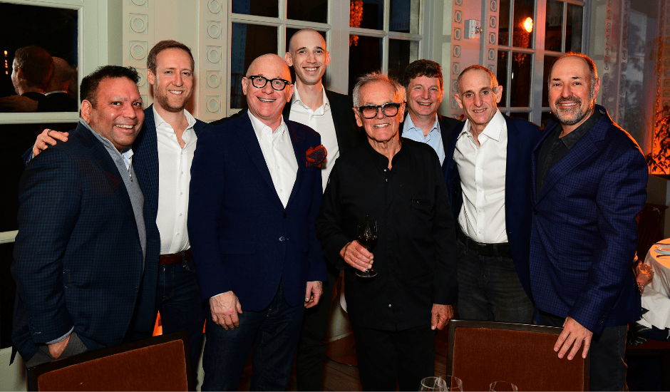 Eight men pose together smiling at a formal indoor dinner with tables and chairs around them.