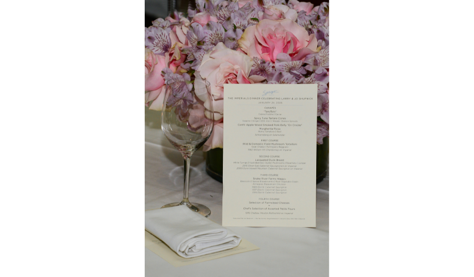 Elegant table setting with a printed dinner menu, folded white napkin, wine glass and pink floral centerpiece.