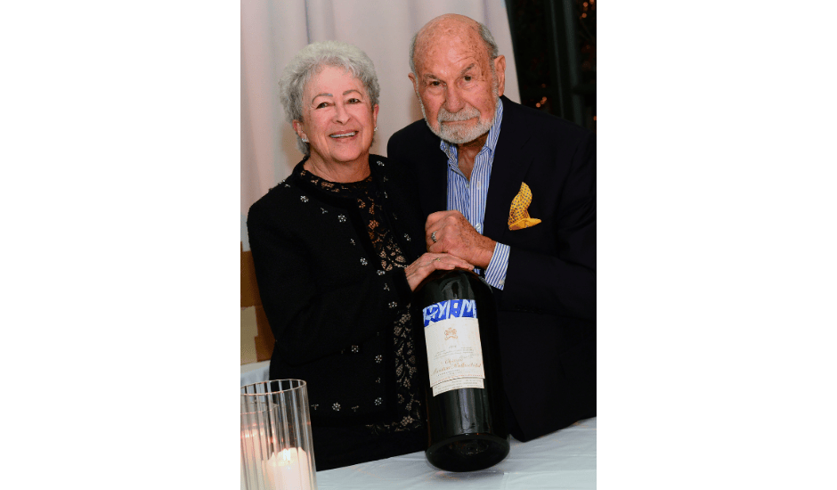 Two adults smile while holding a large wine bottle at a table during an indoor celebration.