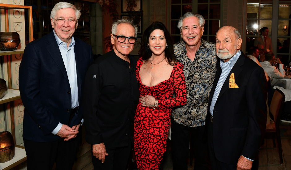 Five adults pose together at an indoor evening event with warm lighting and framed artwork in the background.