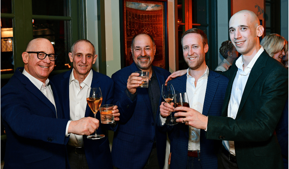 Five men stand close together raising glasses in a toast at an indoor evening reception.