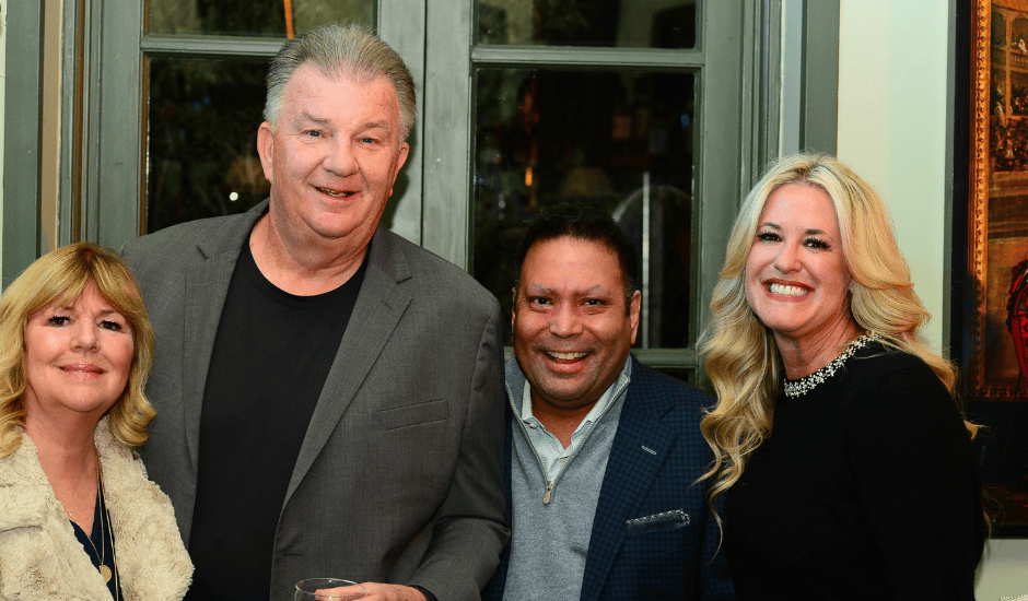 Four adults smile while standing together at an indoor evening event in front of tall windows.