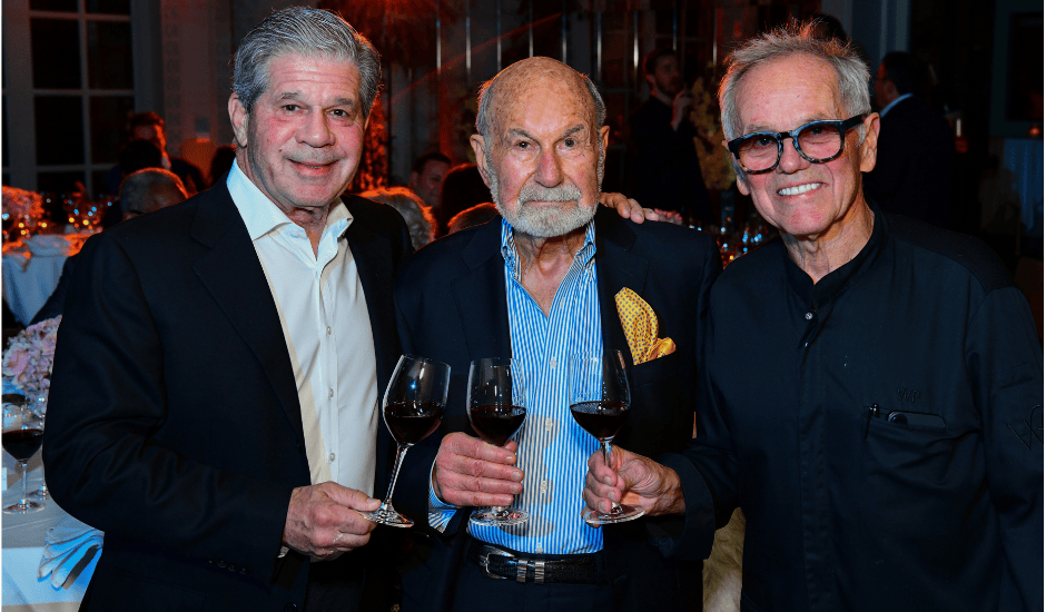 Three men stand close together holding wine glasses at a formal dinner with softly lit tables behind them.
