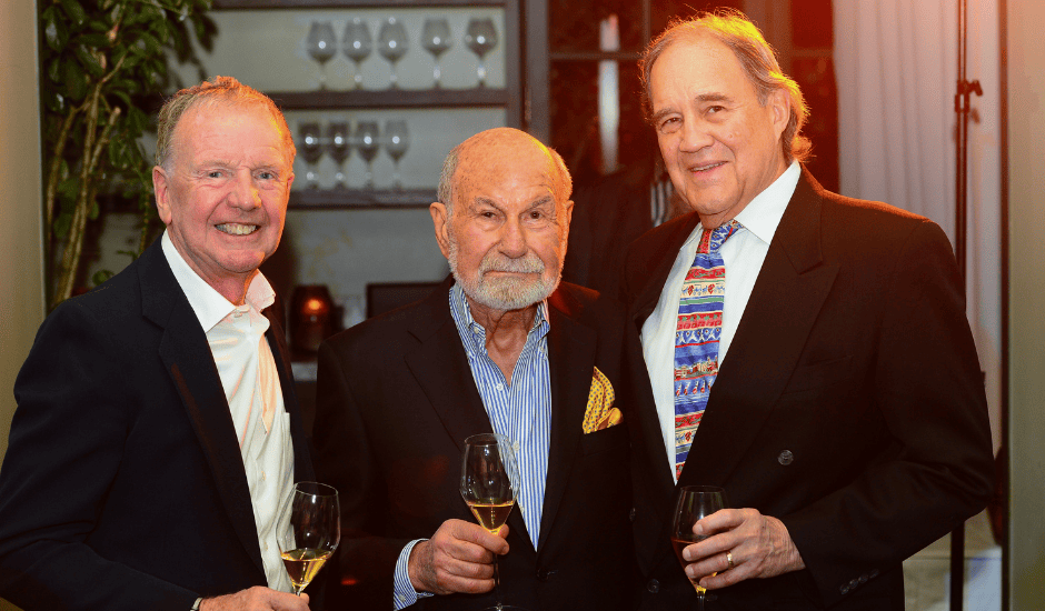 Three men pose with wine glasses at an indoor reception with shelves of glassware in the background.