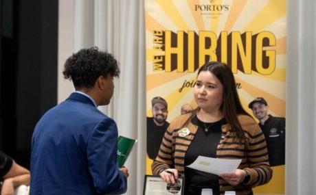 Woman standing a table speaking to a student in professional attire in front of a Porto's hiring banner.