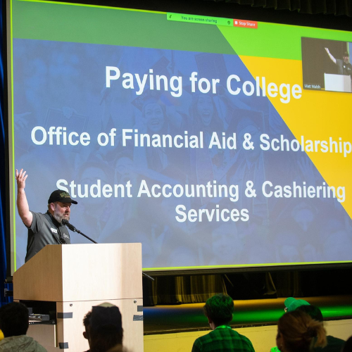 A man giving a presentation in front of a screen which reads "Paying for College, Office of Financial Aid & Scholarships, Student Accounting & Cashiering Services"