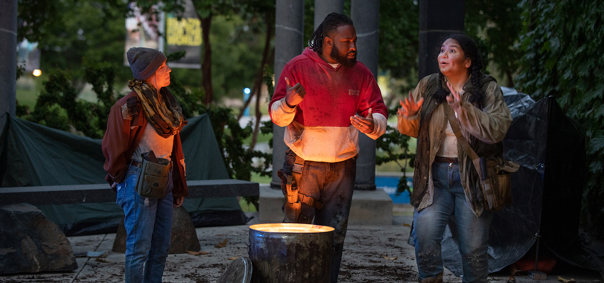 Three cast members in a scene around a trash can fire pit
