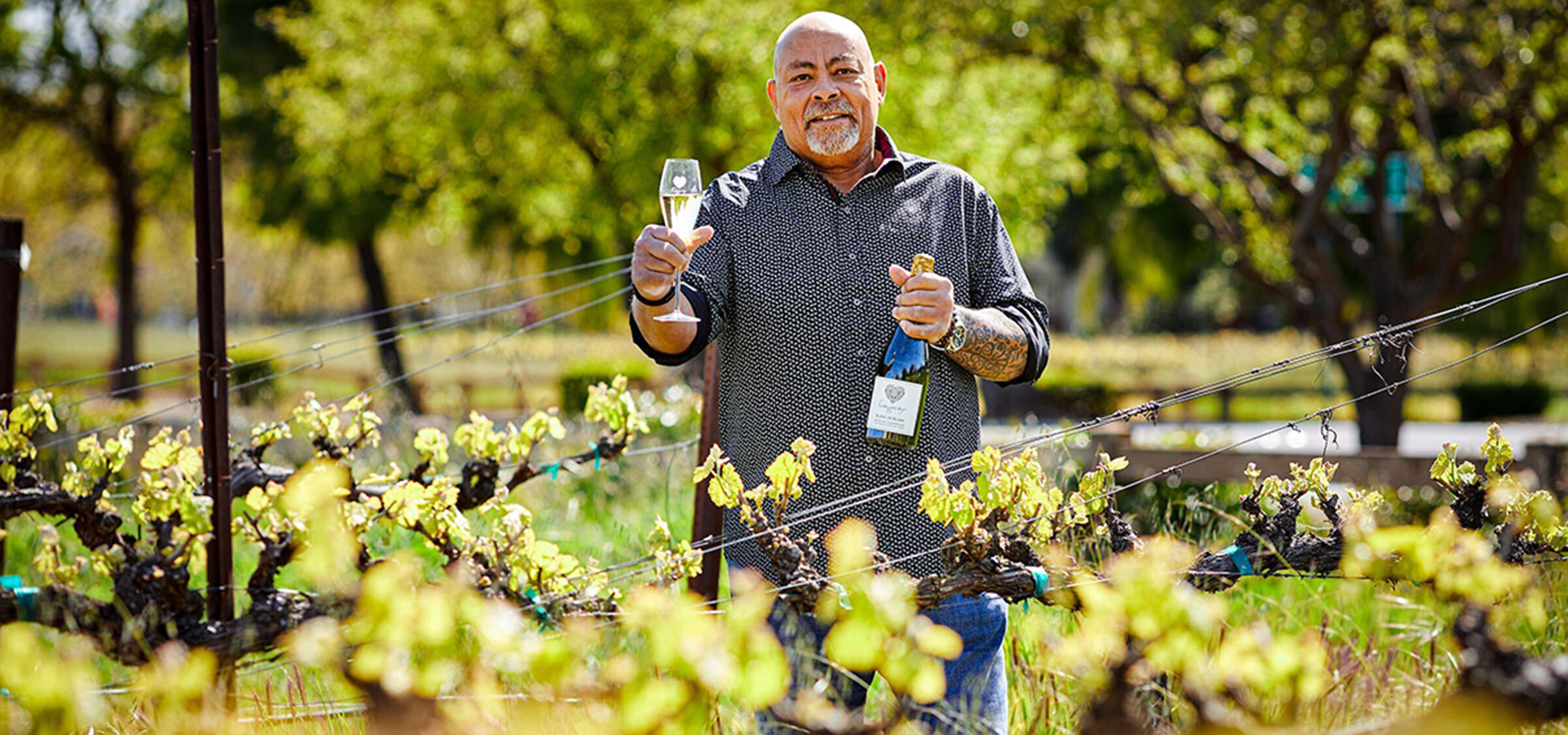 Phil Long holding a bottle and glass of his signature wine while standing in the vinyard
