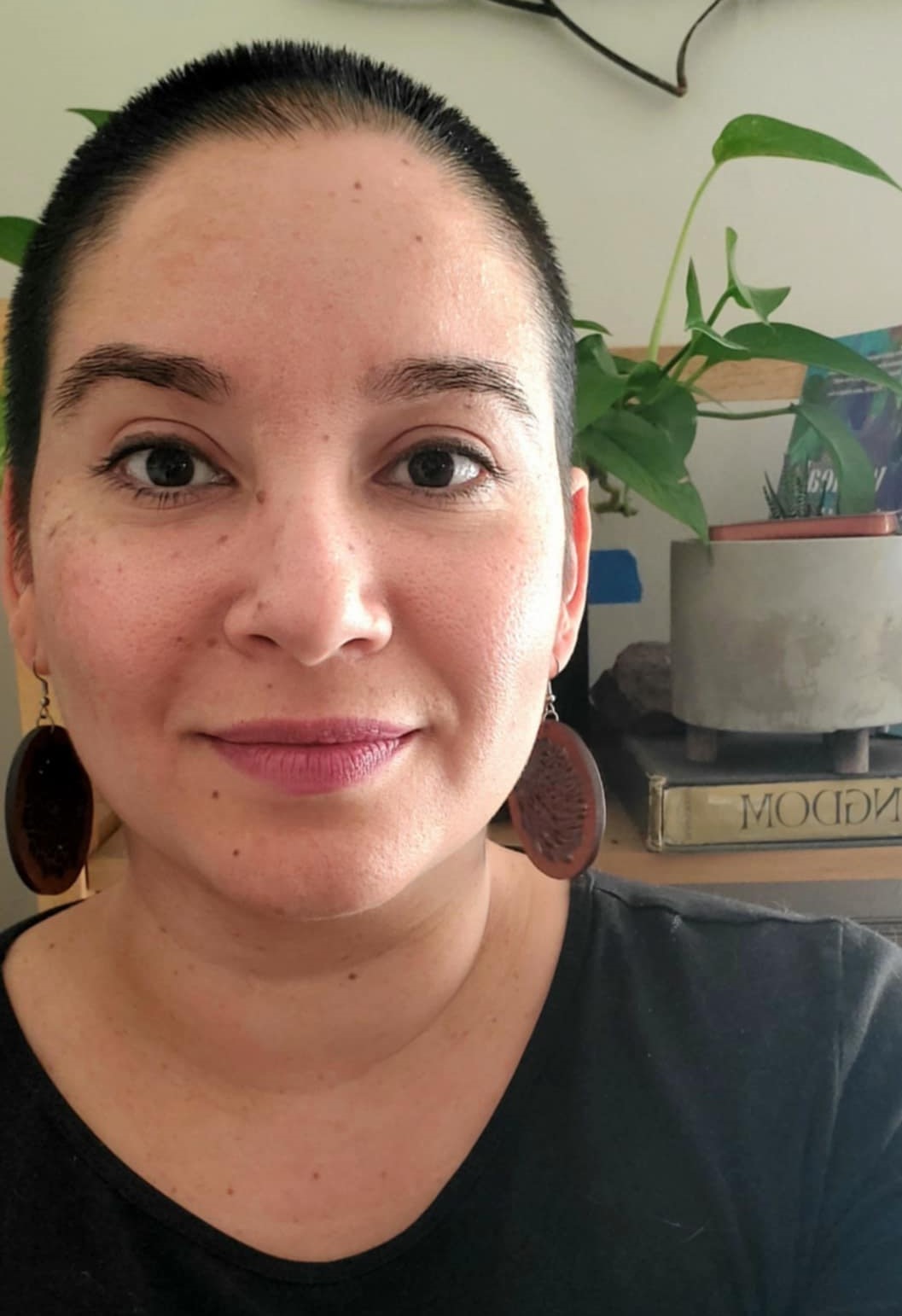 An olive-skin-toned femme with buzzed hair, a black shirt, large wooden earrings, and pink lipstick smiles at the camera. She stands behind a bookshelf with plants.