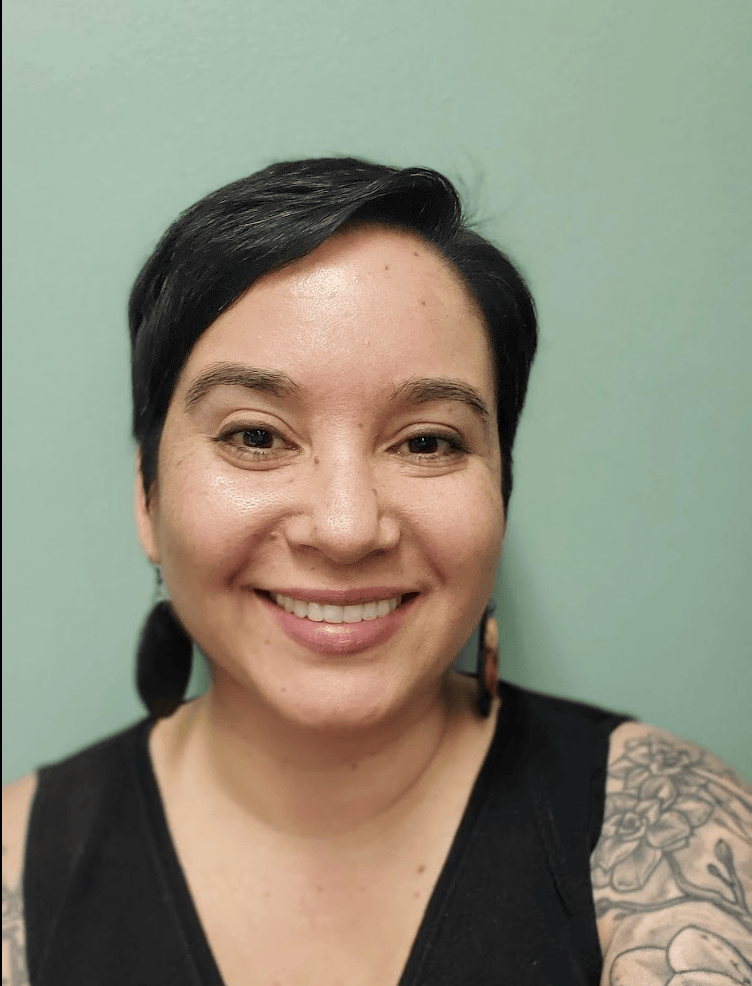 An olive-skin-toned femme with buzzed hair, a black shirt, large wooden earrings, and pink lipstick smiles at the camera. She stands behind a bookshelf with plants.
