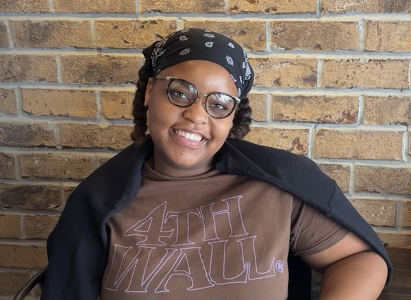 Aliya possing with a bandana on her head, a black sweater over her shoulders and a brick wall behind her.