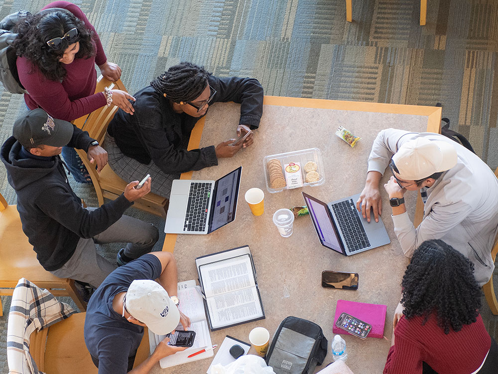 six students studying at a table with laptops and mobile devices
