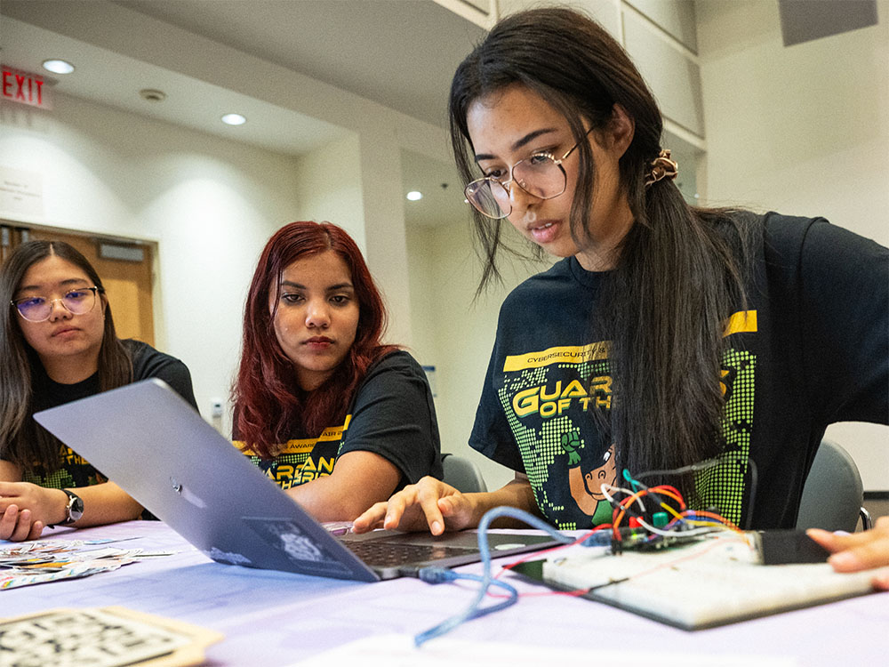 three students seated at a table all looking at one laptop