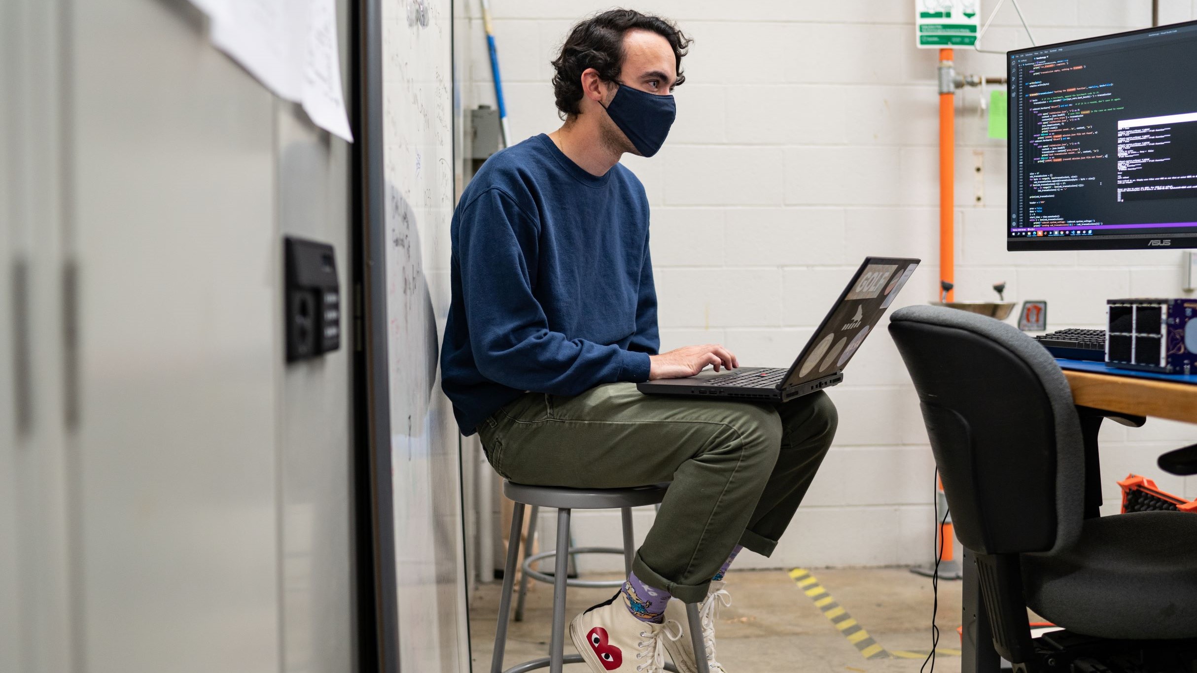 A male engineering student sitting on a stool and working on a project with his laptop.