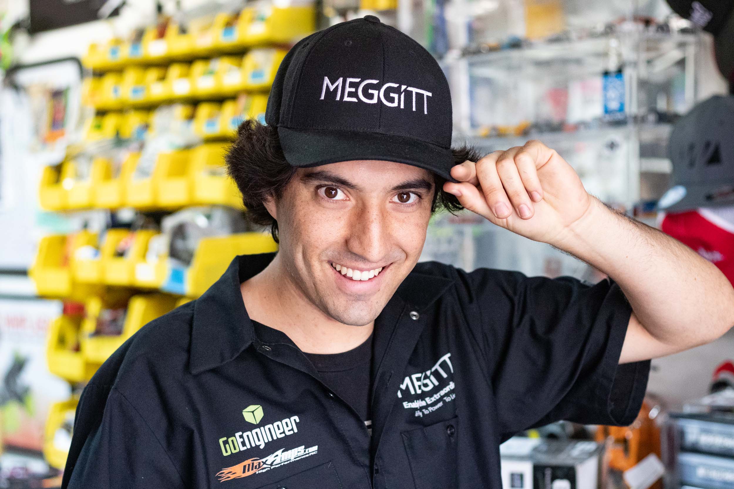 A smiling, male CPP Engineering student tipping his black cap. The cap has the text MEGGITT. His button shirt has logos with the text Go Engineer and Max Amps.