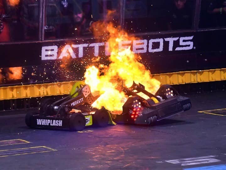 Two human-controlled battle robots engage in combat in an arena. One of the robots is one fire. A robot on the left has the text Whiplash. Battlebots signage is visible behind the robots.