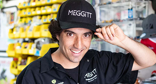 A smiling, male CPP Engineering student tipping his black cap. The cap has the text MEGGITT. His button shirt has logos with the text Go Engineer and Max Amps. 