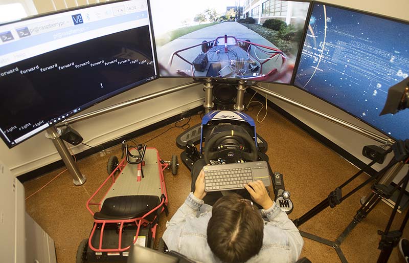 An engineering student in front of three monitors joined together. Student uses a keyboard to remote control a kart.