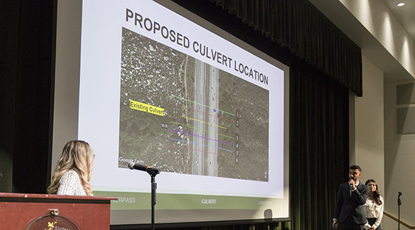 A presentation by three engineering students. A PowerPoint they're using has the title proposed culvert location.